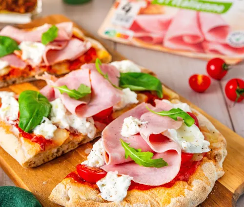 Image d'une pinsa romana, recette italienne avec de la mozzarella, du jambon de porc Fleury Michon et des feuilles de basilique posé sur une pâte type pizza. Le décor est composé d'un paquet de Jambon tranché fin fleury Michon, d'une planche à pain, de tomate cerise et d'accessoires pour manger