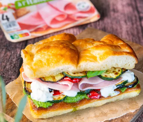 sandwich focaccia, jambon, burrata crémeuse, courgettes grillées, poivrons rouges confits, roquette, pesto, pain doré, texture croustillante, cœur fondant, cuisine méditerranéenne, planche en bois, lumière naturelle, ambiance chaleureuse, food gourmand